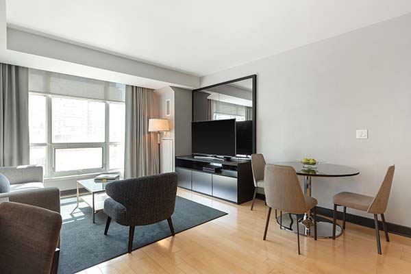 A hotel room featuring two gray chairs, a round dining table with two chairs, and a large flat-screen TV against a wall. Large windows provide natural light, complemented by light-colored wooden flooring.